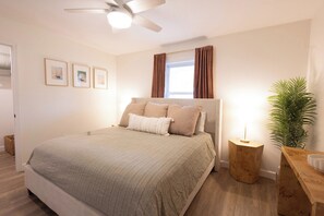 Un dormitorio con cama, ventilador de techo, ventana con cortinas, mesita de noche con lámpara y una planta en maceta.