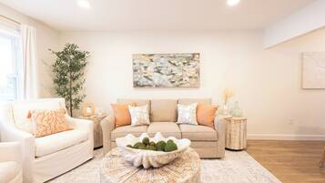 A living room with a beige sofa, white armchairs, a coffee table with decorative items, and a large abstract painting on the wall.