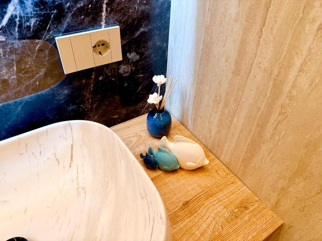 Un bagno con una parete in marmo, un lavandino bianco, un vassoio di legno con oggetti decorativi e un vaso blu con fiori.