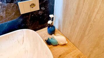Un bagno con una parete in marmo, un lavandino bianco, un vassoio di legno con oggetti decorativi e un vaso blu con fiori.