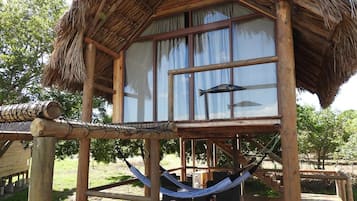 Una estructura de madera con techo de paja, una hamaca y una ventana con cortinas.