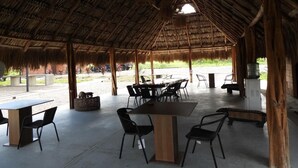 Un comedor al aire libre con mesas y sillas, techo de paja y vistas al exterior.