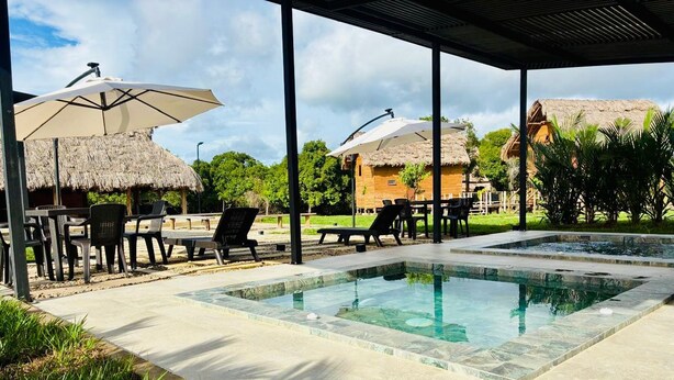 Un área de piscina con sillones de descanso y sombrillas, rodeada de construcciones con techo de paja.
