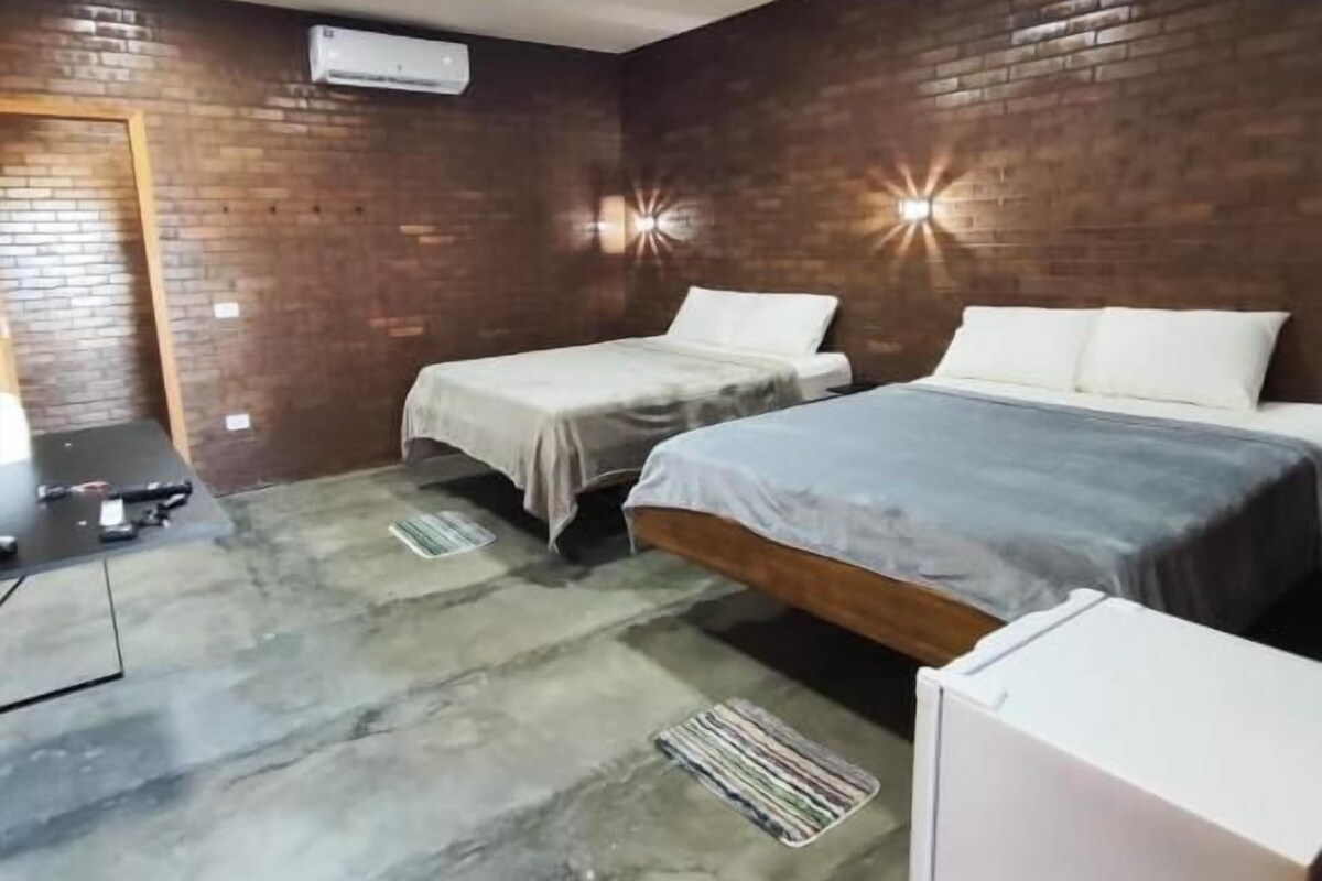 Une chambre avec deux lits, un mur en briques, un ventilateur de plafond et une petite armoire blanche.