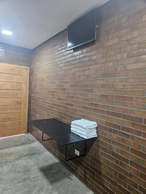 A room with a brick wall, a wooden door, a black table with towels, and a mounted TV. - Hoteis Online  (Boa Vista)