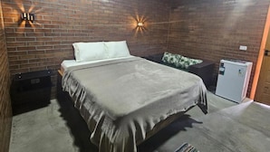 Un dormitorio con pared de ladrillo, una cama, una mesita de noche, un refrigerador pequeño y luces fijadas en la pared.