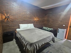 Une chambre avec un mur en briques, un lit, une table de chevet, un petit réfrigérateur et des lampes fixées au mur.
