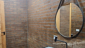 Un baño con pared de ladrillo, puerta de madera, espejo redondo y lavabo blanco sobre encimera negra.