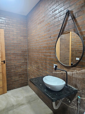 Une salle de bain avec un mur en briques, une porte en bois, un miroir rond et un lavabo blanc doté d’un plan de travail noir.