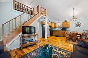 A spacious living area with a wooden staircase, a flat-screen TV, a dining table with chairs, and a kitchen with wooden cabinets. - Gorge-Us Getaway: A Cozy Retreat for Family and Friends (Zoe)