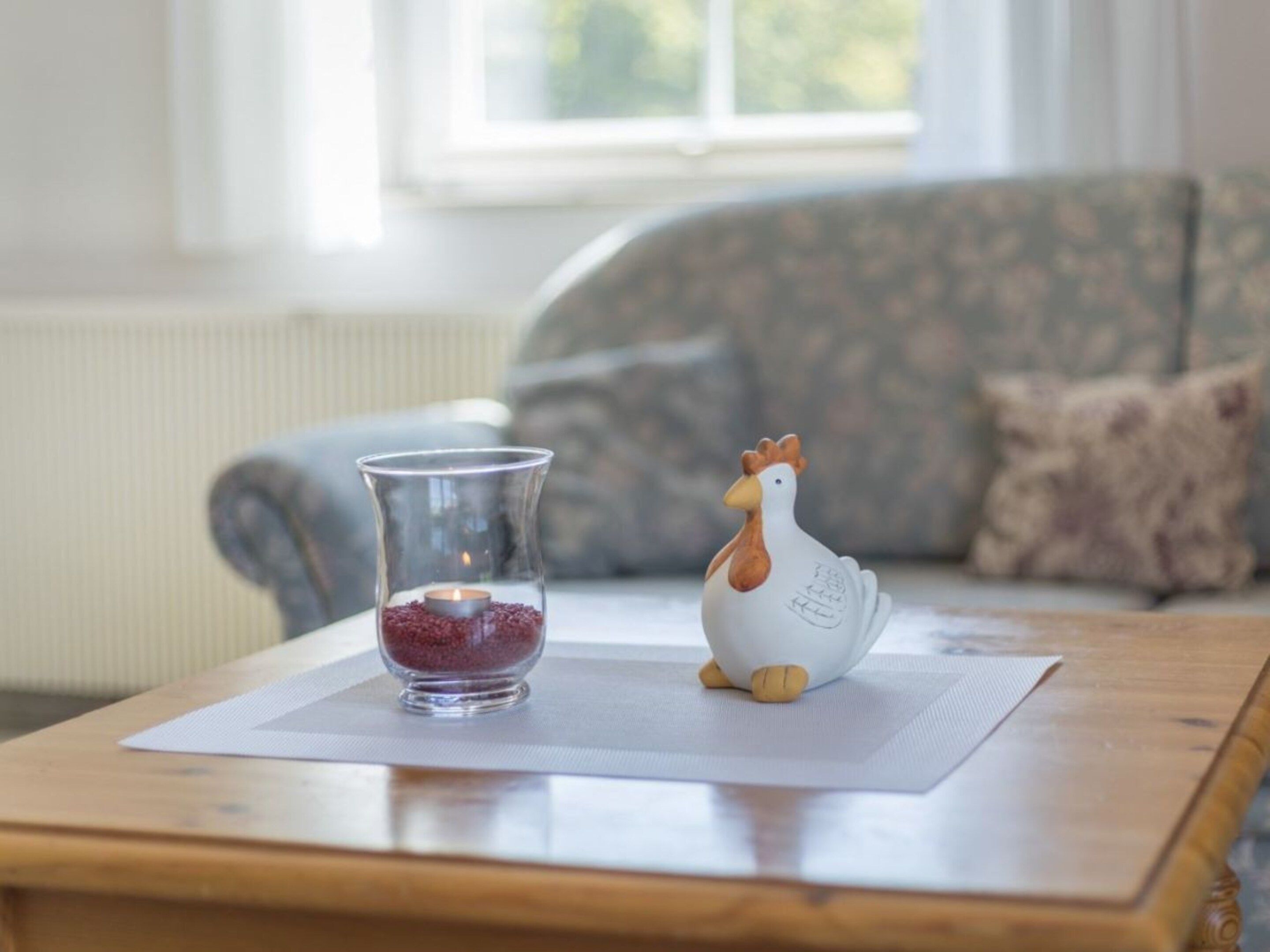 A clear glass candle holder with a lit candle on a wooden table, a ceramic chicken figurine, and a patterned sofa in the background.