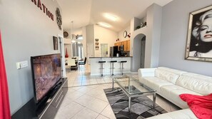 A modern living room with a flat-screen TV, a glass coffee table, a white sofa, and a kitchen area in the background. - 4 BD/ 3 BA/ Rolling Hills Estates (7952 HE) (Kissimmee)