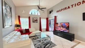 A modern living room with a sofa, a glass coffee table, a flat-screen TV, and a ceiling fan. - 4 BD/ 3 BA/ Rolling Hills Estates (7952 HE) (Kissimmee)