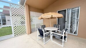 Eine Terrasse mit rundem Tisch und Stühlen, einem Reetdachschirm und Blick auf ein Haus durch eine Gittertür.