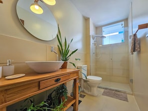 A bathroom with a wooden vanity, a large mirror, a white sink, a toilet, and a shower area. - Barra - Salvador (Salvador)