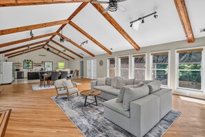 A spacious living area with a high ceiling, exposed wooden beams, a grey sofa, a dining table, and a kitchen in the background.