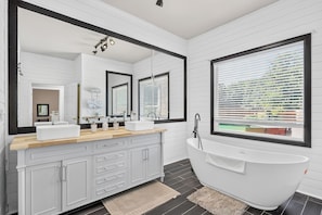 Une salle de bain moderne avec une baignoire sur pieds, un grand miroir et une fenêtre avec des stores.