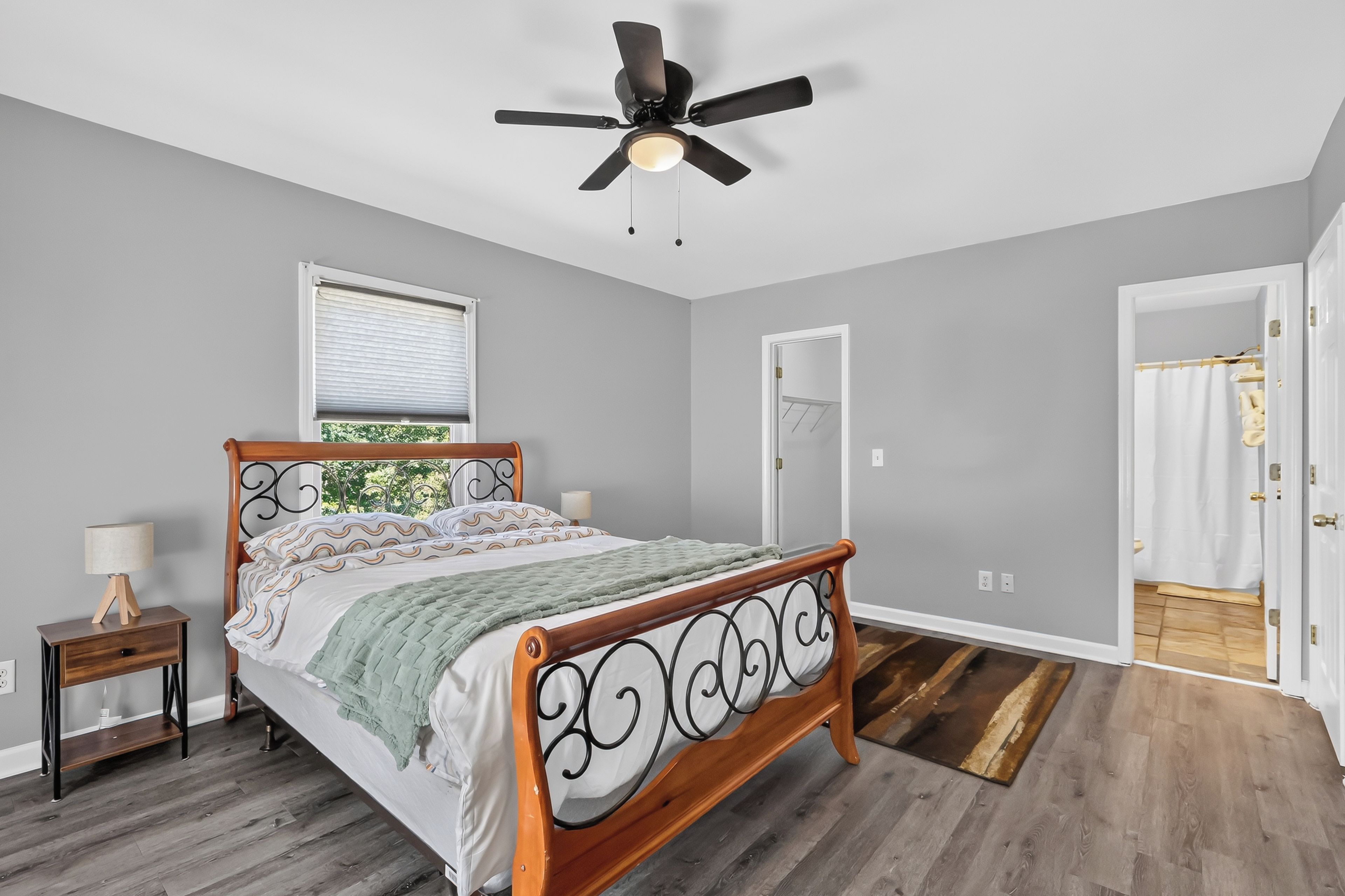 A bedroom with a wooden bed, a ceiling fan, a window with blinds, and a door leading to a bathroom.