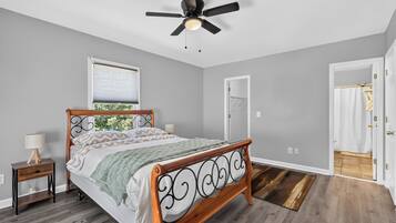 A bedroom with a wooden bed, a ceiling fan, a window with blinds, and a door leading to a bathroom.