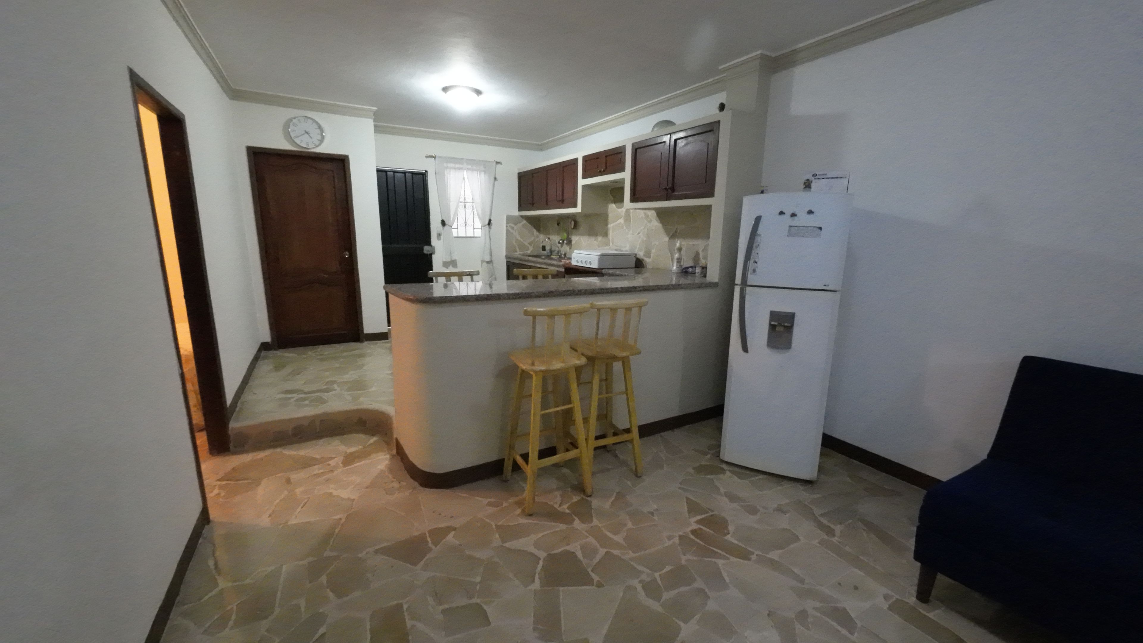 A kitchen with a stone floor, wooden stools, a refrigerator, and a clock on the wall.