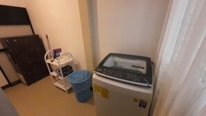A compact room with a refrigerator, a small shelf with cleaning supplies, and a blue laundry basket.