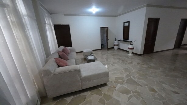 Sala de estar com sofá, mesa de centro e piso ladrilhado. Há portas e uma pequena mesa com objetos decorativos.