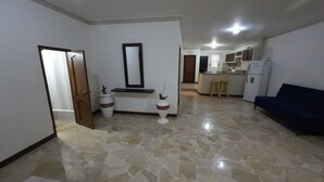 Sala de estar com sofá escuro, área de cozinha com geladeira e pia, e corredor com porta que dá para outro quarto.