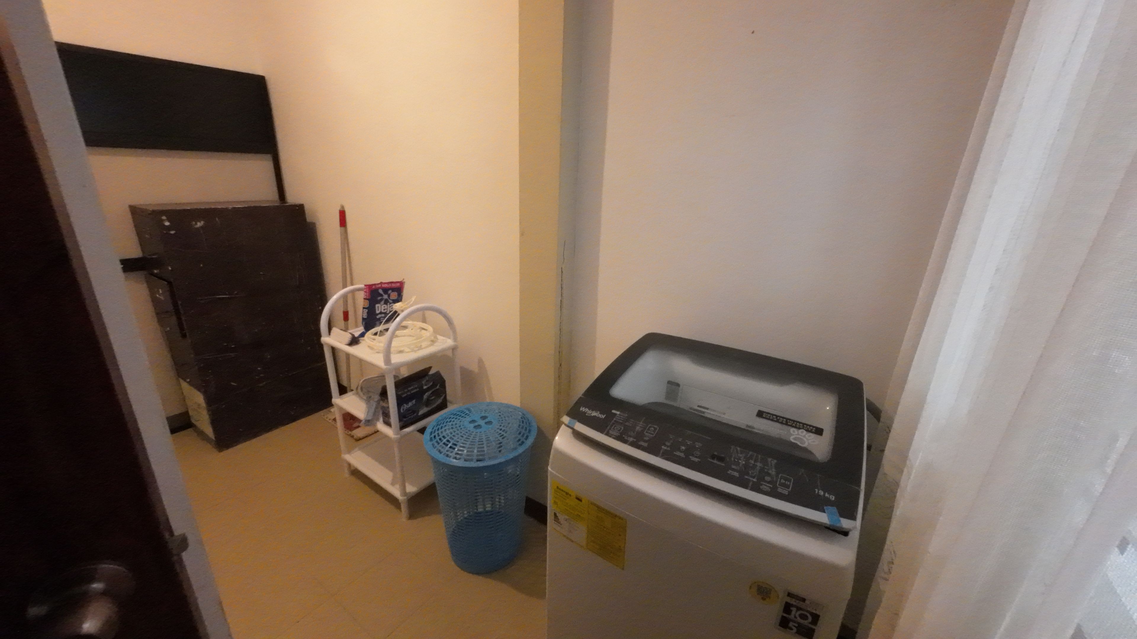 A compact laundry room with a Whirlpool washer, a blue laundry basket, and a shelf with various items.