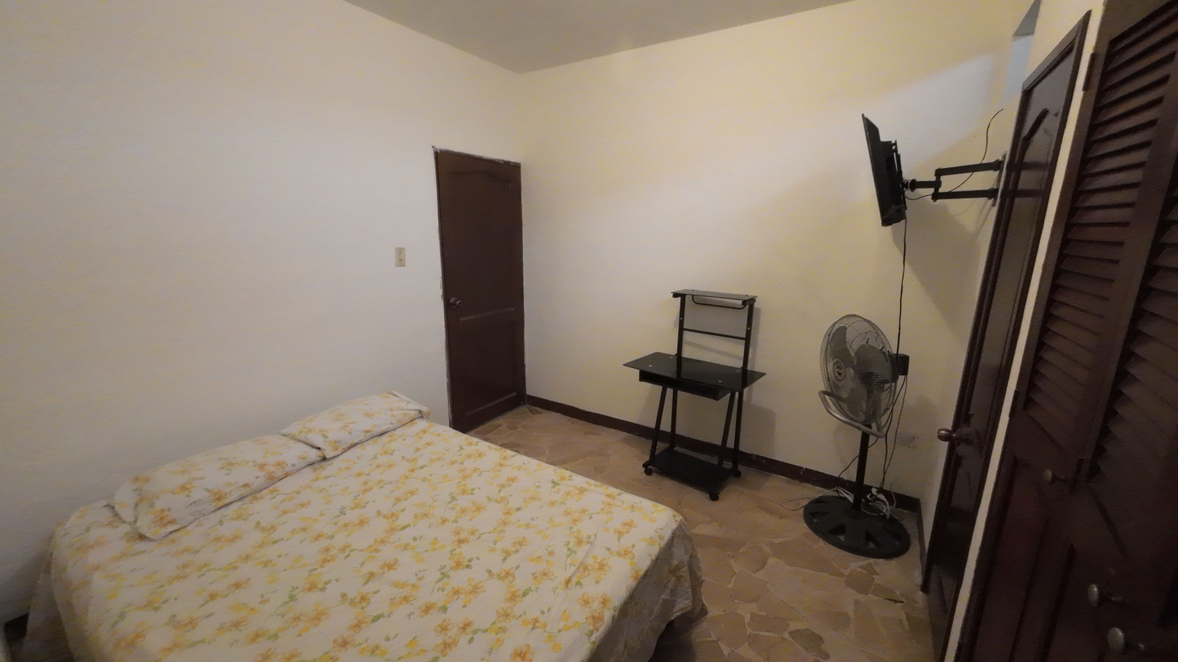 Une chambre à coucher avec un lit, un bureau, une chaise, un ventilateur et une porte.