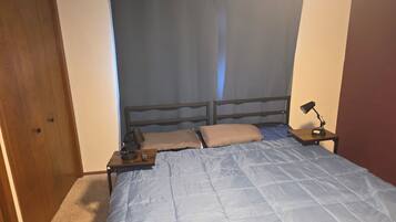 Un dormitorio con cama, mesita de noche, lámpara y cortina.