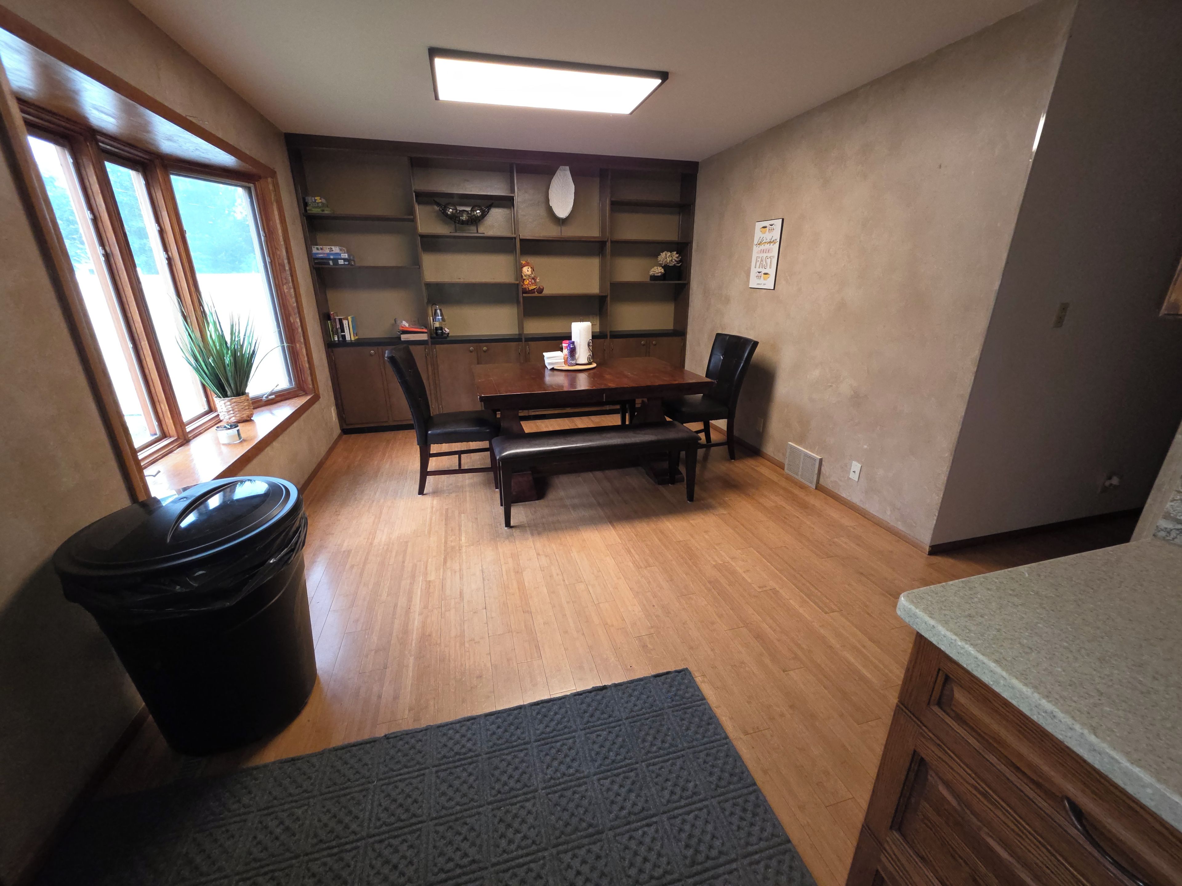 Un comedor con una mesa de madera, sillas, un banco, un cubo de basura y una ventana con vistas.