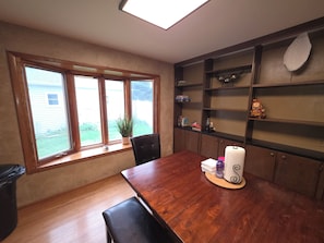 Ein Essbereich mit einem Holztisch, Stühlen, einem Fenster mit Blick auf ein Haus und einem Bücherregal mit diversen Gegenständen.