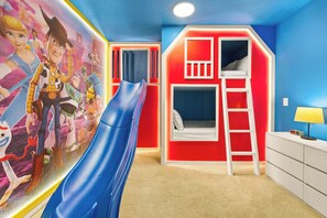 A child's playroom with a blue slide, a wall mural, a bunk bed, and a desk with a lamp. - 2472 Toy Barn Slide Bed & Game Room! Private Pool (Kissimmee)