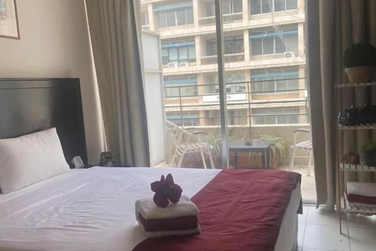 Una habitación de hotel con cama, ventana con cortinas y balcón con vista a un edificio.