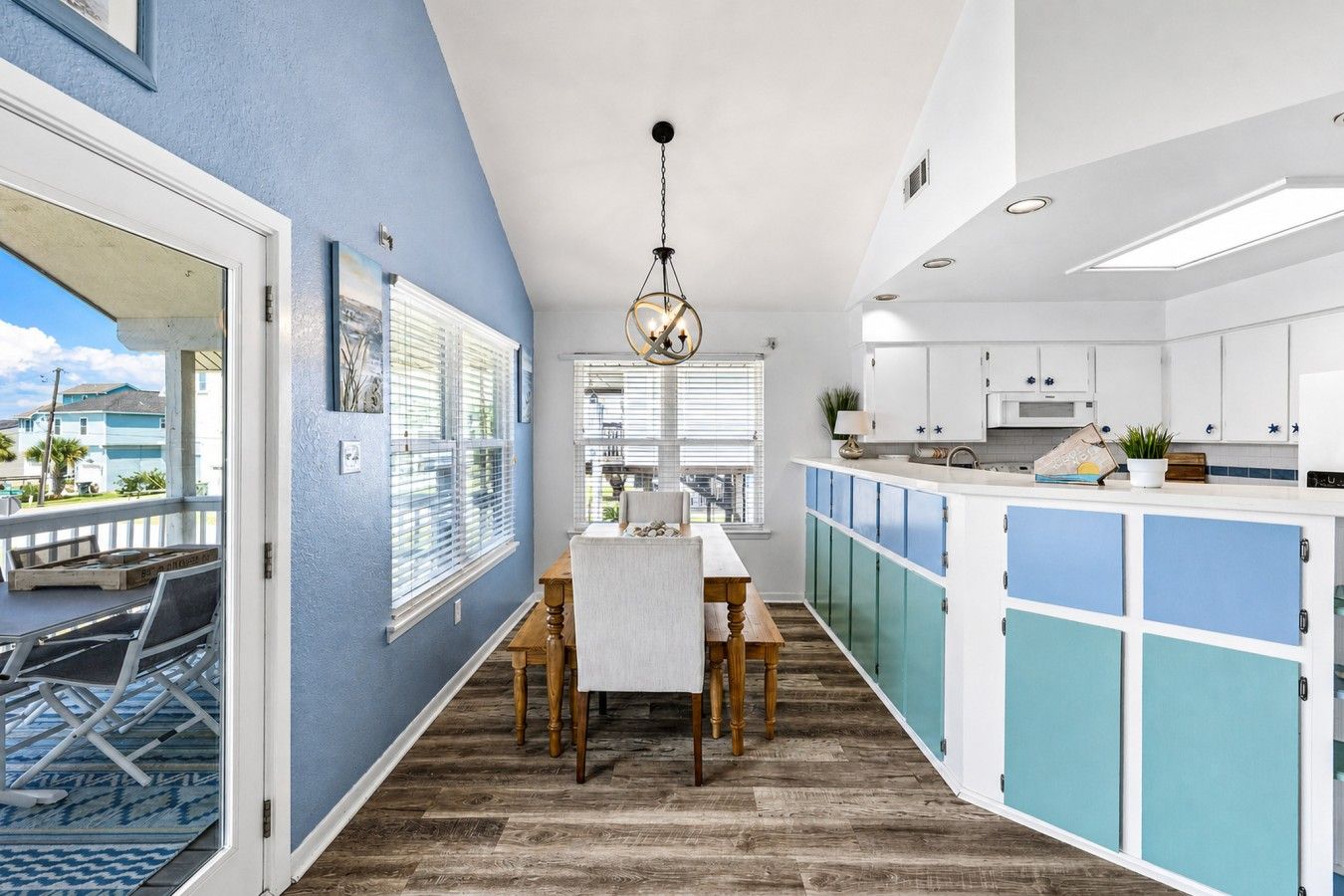 A modern kitchen with a dining area, featuring a wooden table and chairs, a hanging light fixture, and a view through the window.