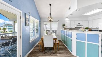 A modern kitchen with a dining area, featuring a wooden table and chairs, a hanging light fixture, and a view through the window.