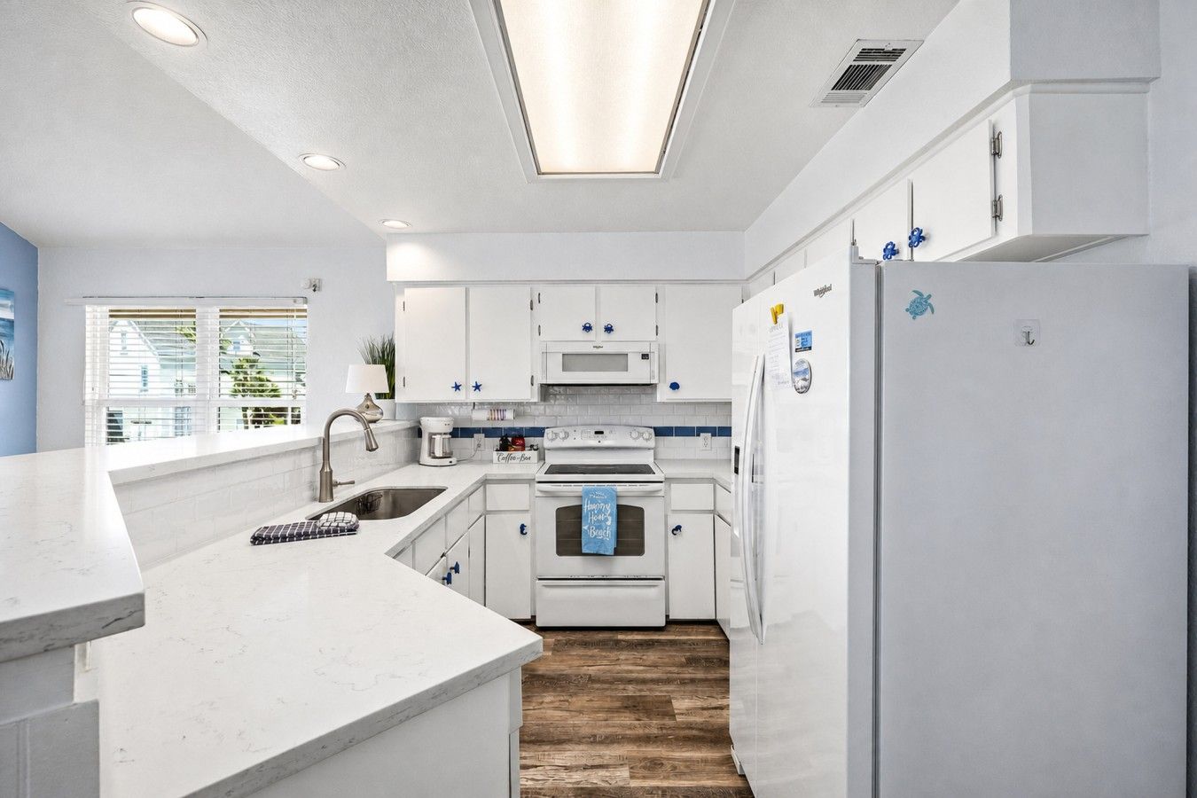 Una cocina moderna con armarios blancos, horno empotrado, refrigerador y ventana con persianas.