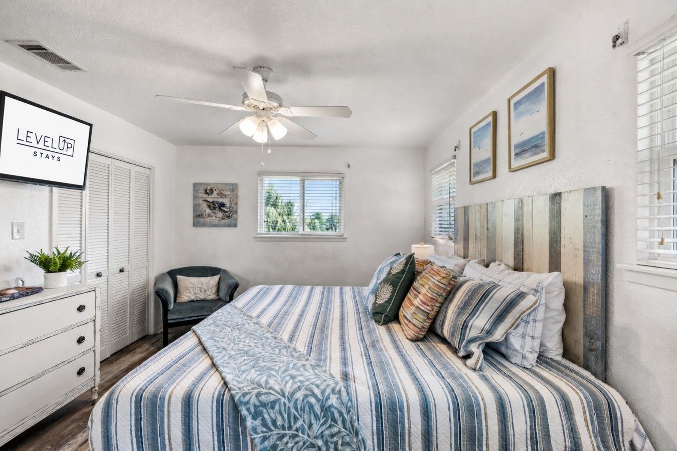 Une chambre à coucher comprenant un lit, un ventilateur de plafond, une fenêtre avec des stores, une table de chevet et une œuvre d’art accrochée au mur.