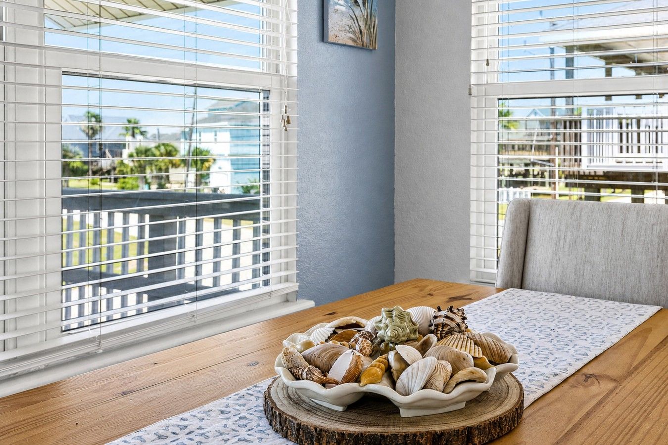 Ein Zimmer mit einem Holztisch, einer Schale Muscheln und Blick durch große Fenster mit Jalousien.