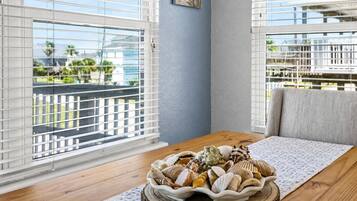 Ein Zimmer mit einem Holztisch, einer Schale Muscheln und Blick durch große Fenster mit Jalousien.
