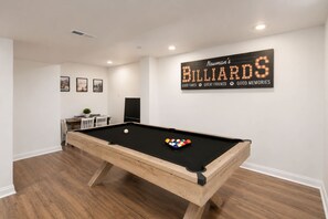 Una mesa de billar en una habitación con un cartel de billar en la pared, suelo de madera y una pequeña zona de estar con una mesa y sillas.