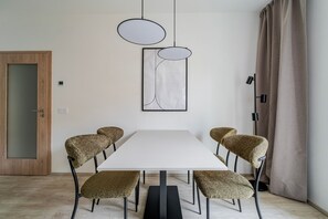 Ein Essbereich mit weißem Tisch, sechs Stühlen, zwei Pendelleuchten, einem gerahmten abstrakten Kunstwerk und einer Holztür.