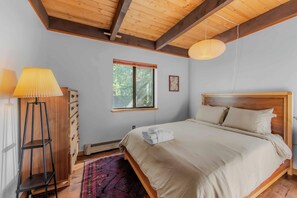 Ein Schlafzimmer mit Holzboden, einem Bett mit weißen Leinen, einer Stehlampe und einem Fenster mit Blick auf Bäume.