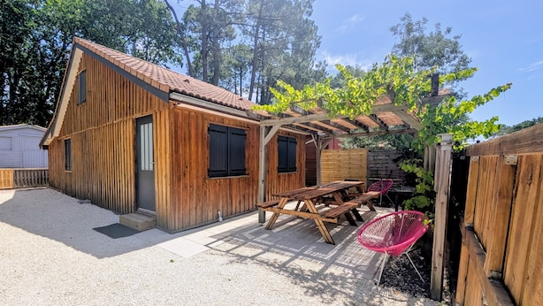A wooden cabin with a covered patio, a picnic table, and a hanging chair. - Chalet in the middle of the woods, ARCACHON BAY (Gujan-Mestras)