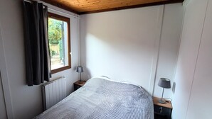 A bedroom with a bed, a window with curtains, a radiator, and a nightstand with a lamp. - Chalet in the middle of the woods, ARCACHON BAY (Gujan-Mestras)