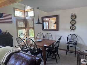 A dining area with a wooden table and chairs, a leather couch, a large window with blinds, an American flag, and wall-mounted decor.