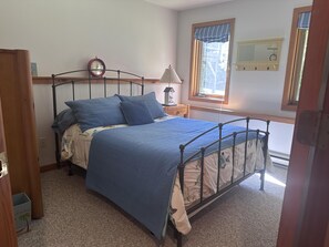 A bedroom with a metal bed frame, a bedside table with a lamp, a clock, and a window with a view.