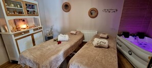 A spa room with two massage tables, a massage chair, a bathtub with water jets, and a shelf with various bottles and a lamp.