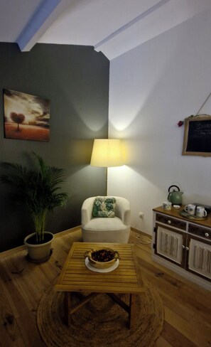A cozy living room with a white armchair, a wooden coffee table with a bowl, a potted plant, a lamp, and a cabinet with kitchen items.