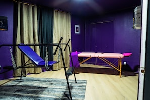 Um quarto com parede roxa, um banco de exercícios azul, uma mesa de massagem rosa e piso de madeira.